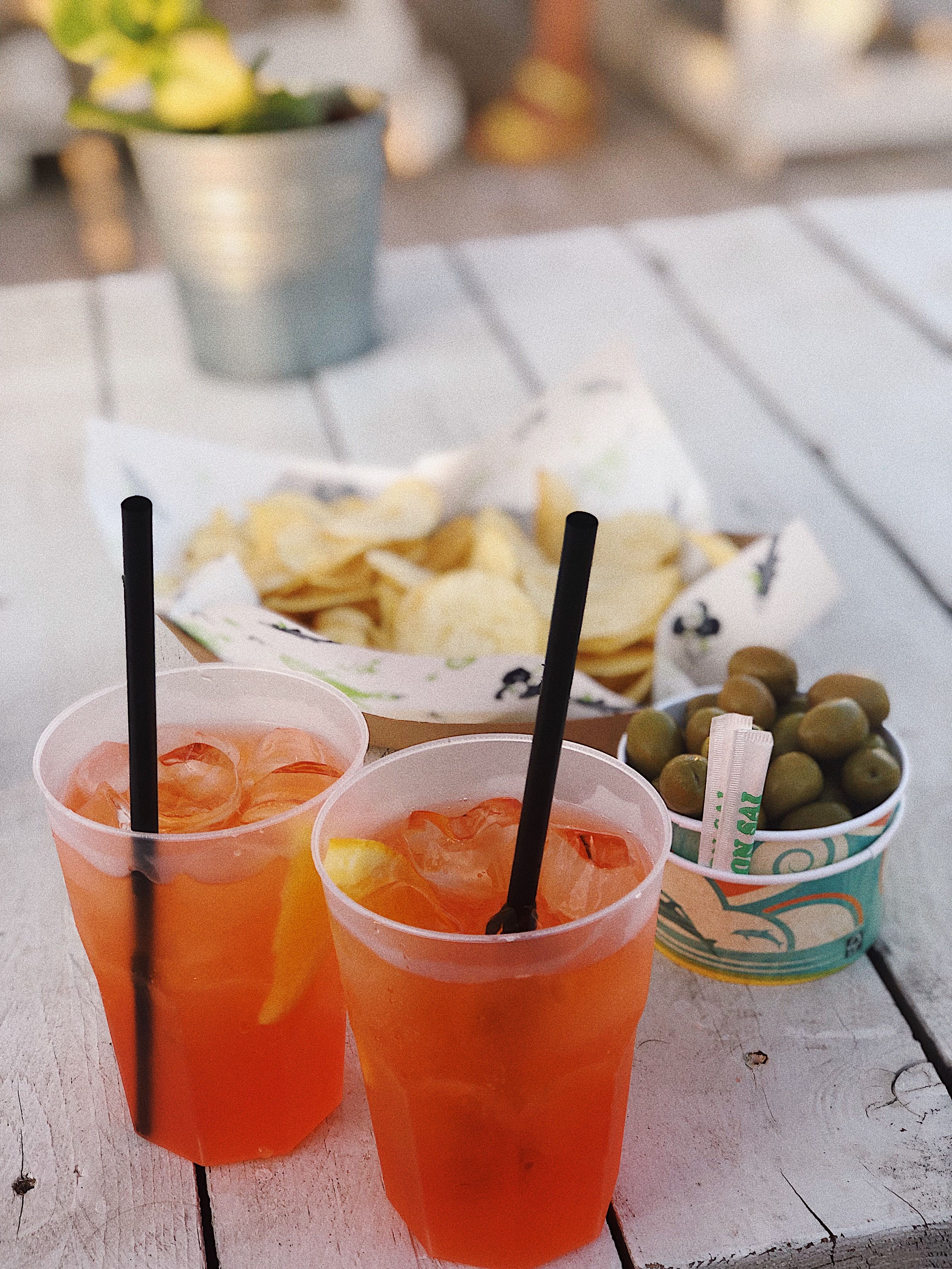 Dwa kubki z pomarańczowym, włoskim aperitifem z lodem i rurkami, serwowane na białym drewnianym stole w towarzystwie zielonych oliwek i chipsów.