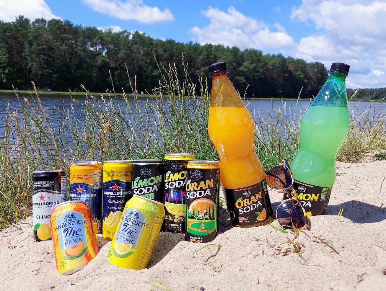 Włoskie napoje gazowane w puszkach i butelkach, w tym Lemon Soda, Oran Soda i San Pellegrino, ułożone w piasku na plaży na tle jeziora i lasu.